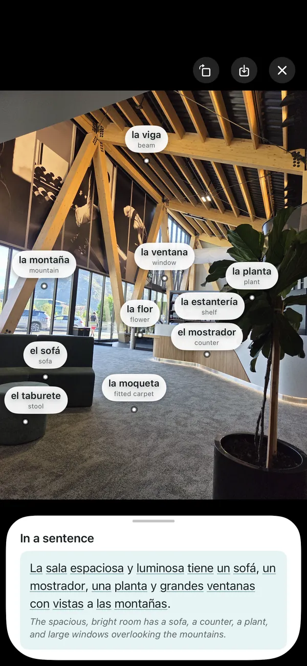 A photo of a bright room with Spanish labels on the things in it, such as la ventana for window and el sofá for sofa, above a Spanish sentence describing the room with its English translation